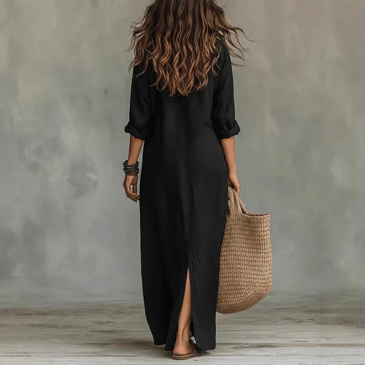 Back view of a woman wearing a black linen maxi shirt dress with a side slit, styled with a woven straw tote bag.
