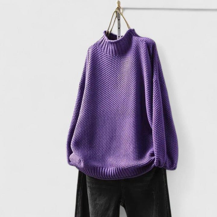 Purple textured mock neck knitted jumper with dropped shoulders and ribbed trim, hanging against a white wall.