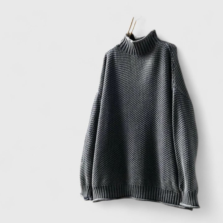 Charcoal grey mock neck knitted jumper with a textured finish, dropped shoulders, and ribbed hem hanging against a wall.