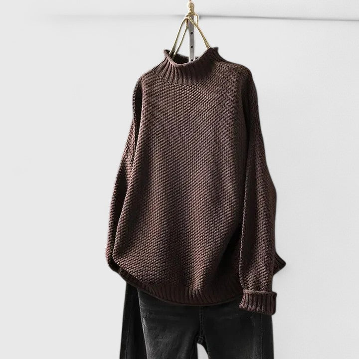 Dark brown mock neck jumper in a textured knit with dropped shoulders and ribbed cuffs, hanging against a white wall.