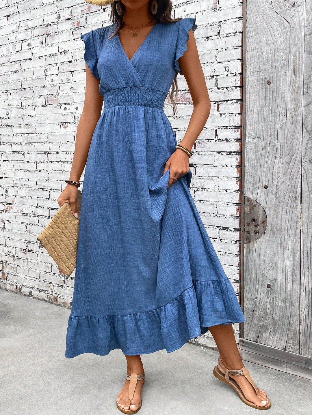Blue V-neck maxi dress with flutter sleeves, a smocked waist, and a tiered ruffle hem against a white brick wall.