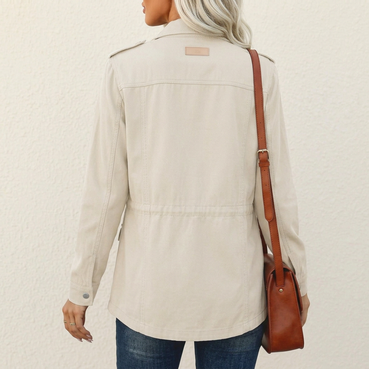 Back view of a beige utility jacket with shoulder tabs, a cinched waist, and a brown crossbody bag.