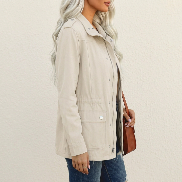 Beige utility jacket with shoulder tabs, flap pockets, and a full-length zip closure worn over blue denim jeans.
