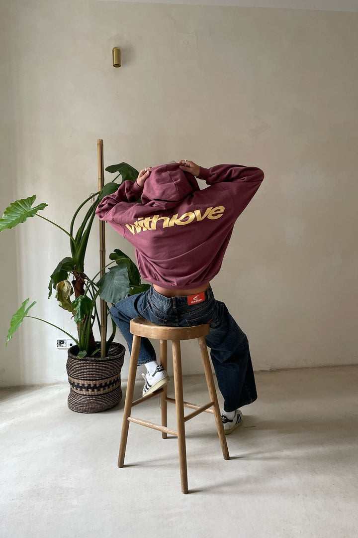 Oversized plum-colored pullover hoodie with gold back lettering, worn with baggy blue jeans while sitting on a wooden stool.