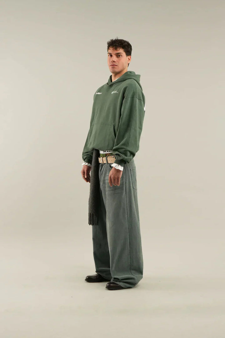 Model wearing an oversized sage green pullover hoodie with a front kangaroo pocket and subtle white logo detailing.