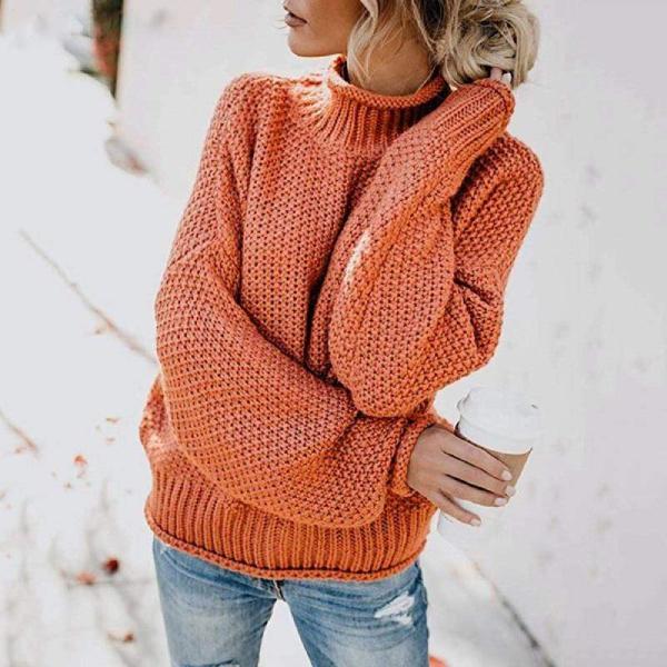 Woman wearing a chunky knit orange turtleneck jumper with dropped sleeves and ribbed cuffs paired with blue jeans.