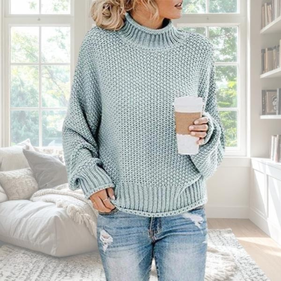 Woman wearing a sage green chunky knit turtleneck jumper with dropped sleeves and ribbed trim, styled with blue jeans.