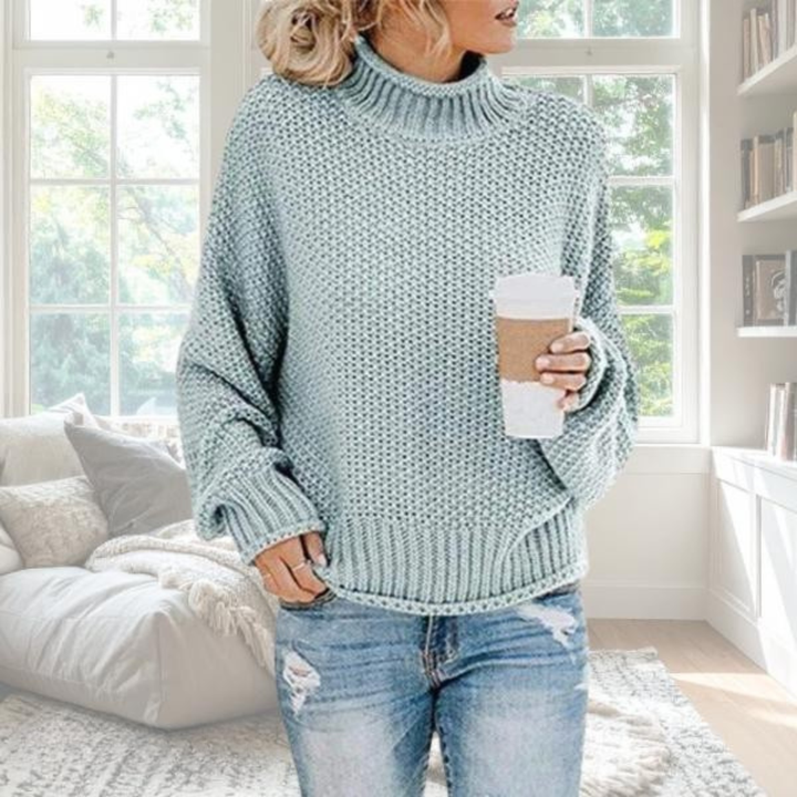 Woman wearing a sage green chunky knit turtleneck jumper with dropped sleeves and ribbed trim, styled with blue jeans.