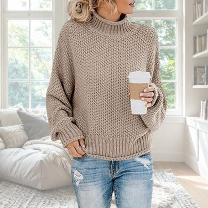 Woman wearing a beige chunky knit turtleneck jumper with dropped sleeves and ribbed trim, styled with blue jeans.