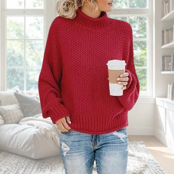 Woman wearing a red chunky knit turtleneck jumper with dropped sleeves and ribbed cuffs in a cosy indoor setting.