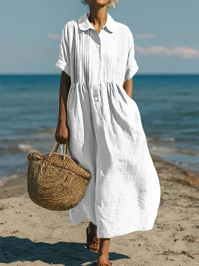 White linen-look maxi shirt dress with pintuck bodice, button placket, and short sleeves, worn on a sunny beach.