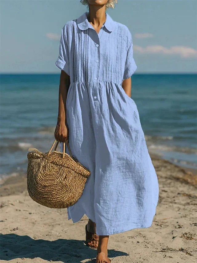 Light blue linen-look maxi shirt dress with pintuck bodice, button placket, and short sleeves on a beach.