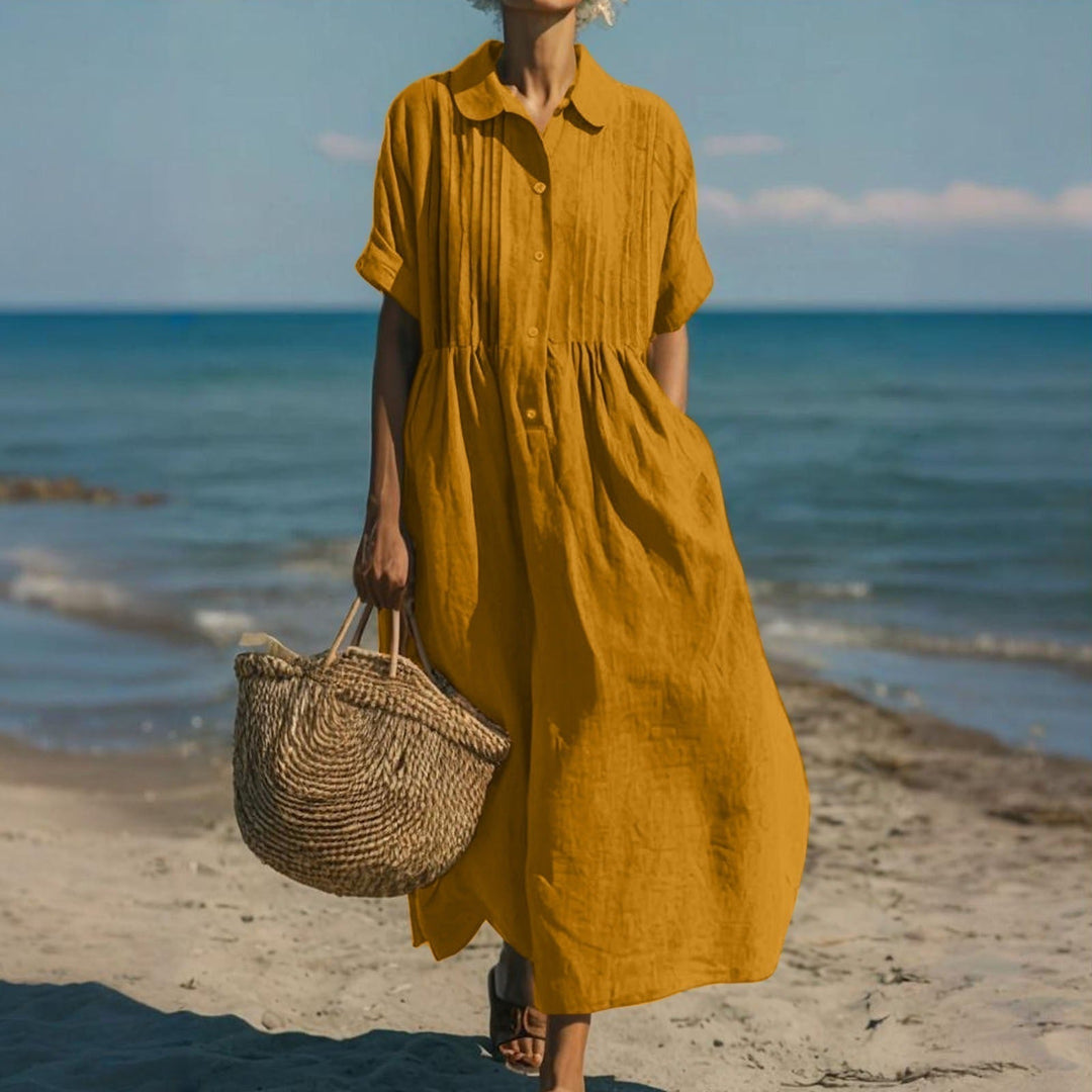Mustard yellow linen-look maxi shirt dress with pintuck bodice, button placket, and short sleeves on a beach.
