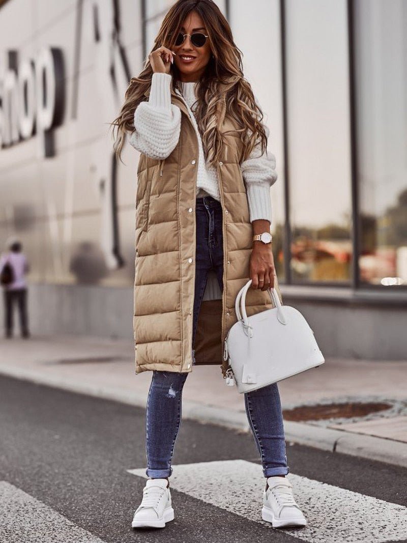 Woman wearing a beige longline padded hooded gilet over a white sweater, blue jeans, and white sneakers.