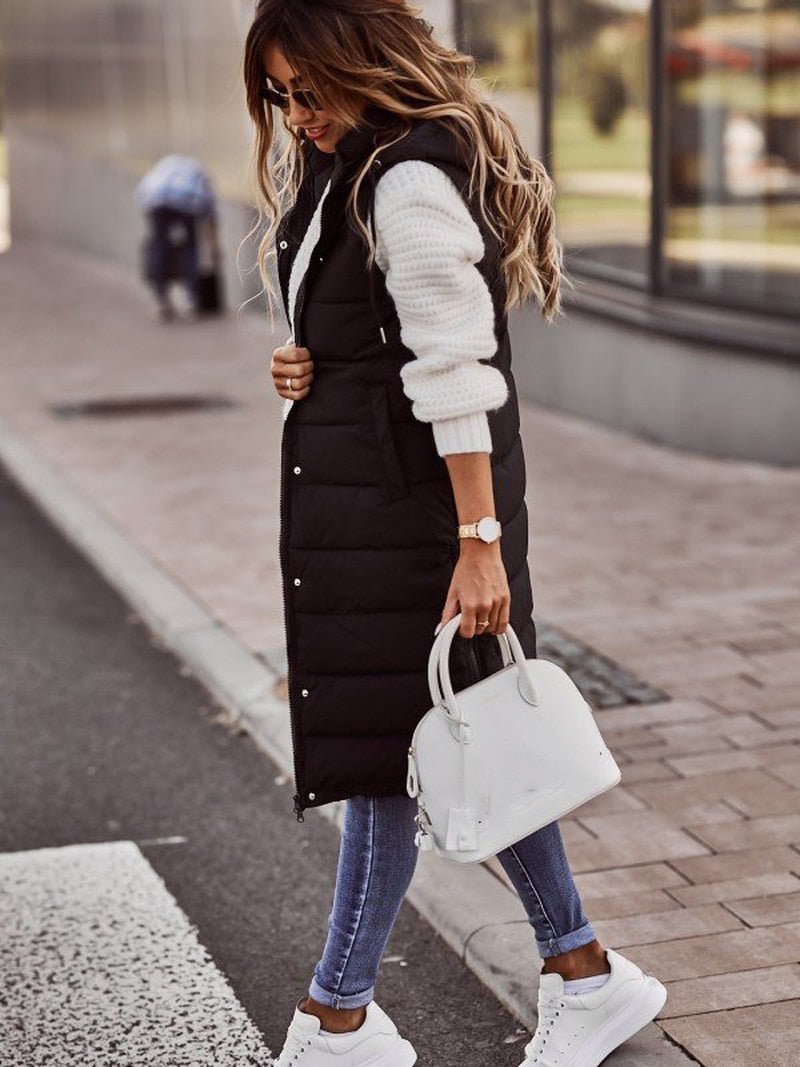 Woman wearing a black longline padded hooded gilet over a white sweater, blue jeans, and white trainers.