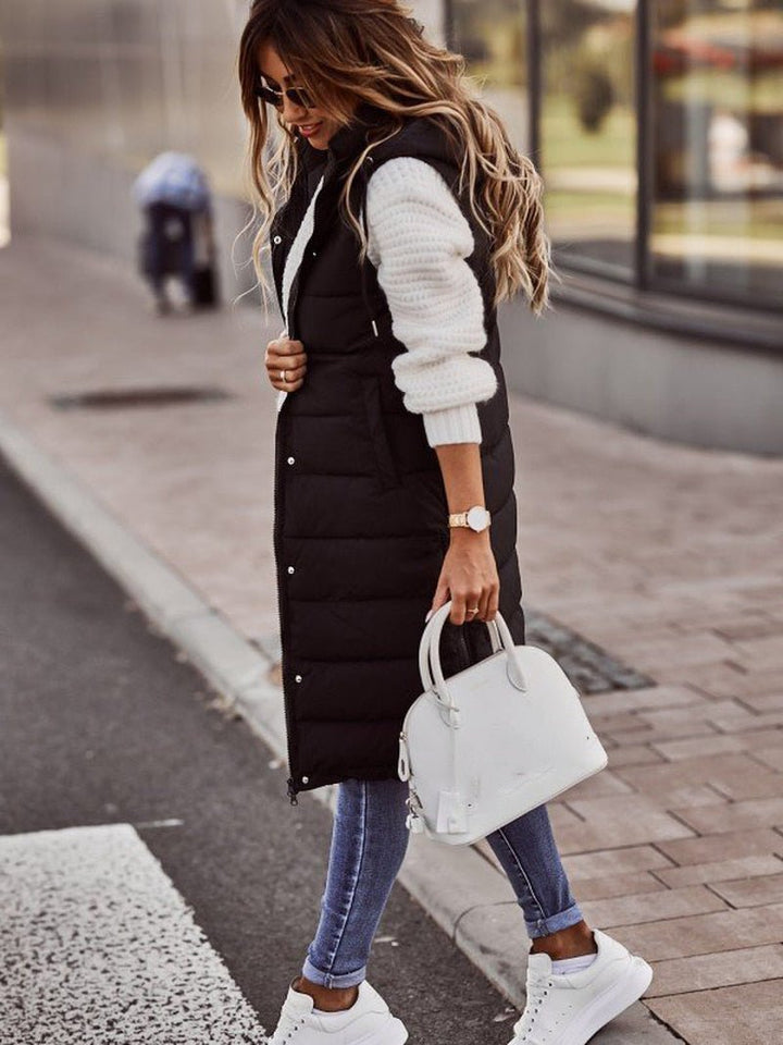 Woman wearing a black longline padded hooded gilet over a white sweater, blue jeans, and white trainers.