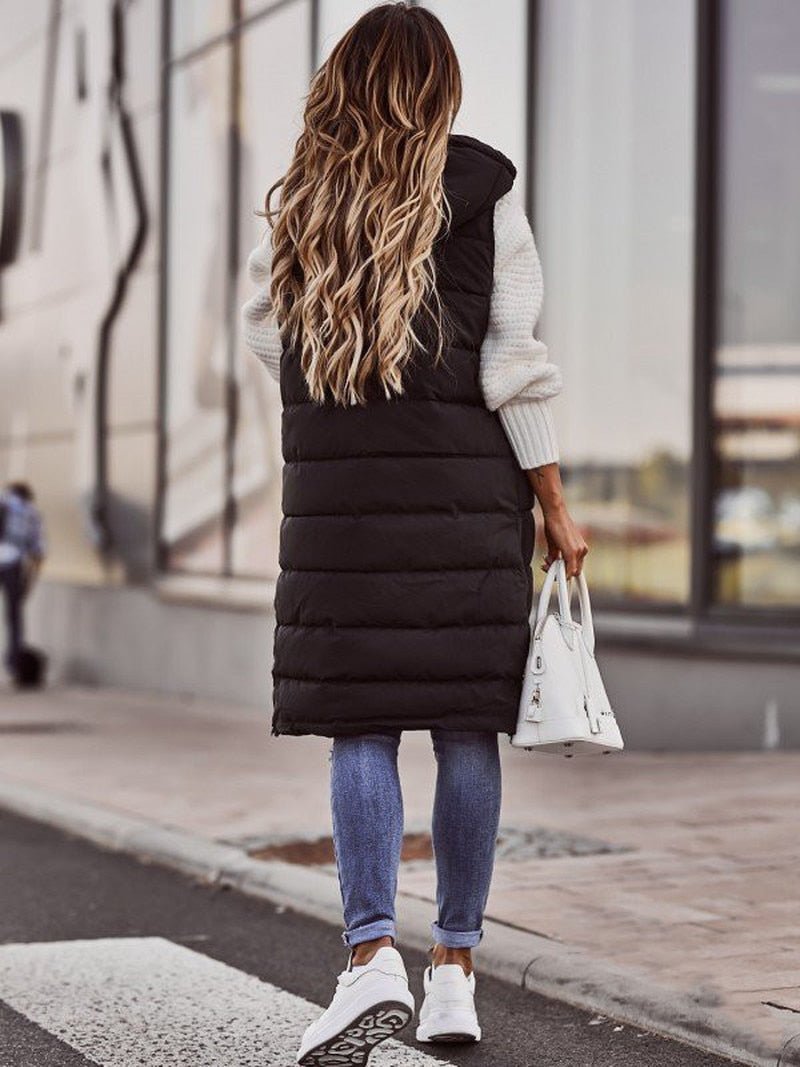 Woman wearing a black longline padded hooded gilet over a white knit sweater, blue jeans, and white sneakers.