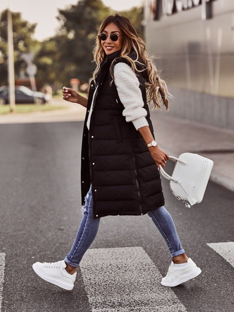 Woman wearing a black longline padded hooded gilet over a white sweater, blue jeans, and white sneakers.