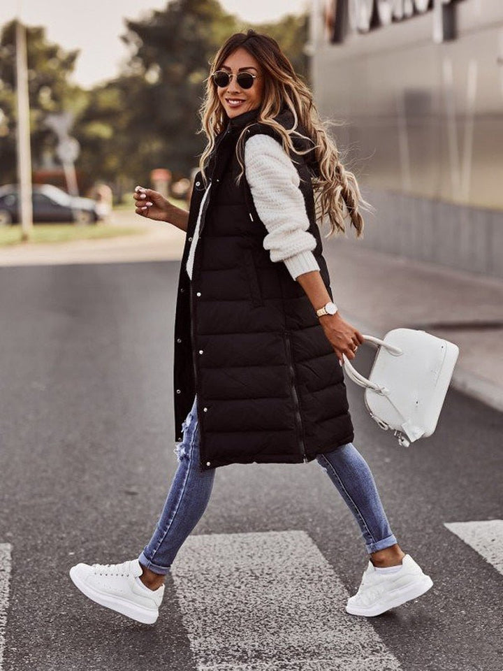 Woman wearing a black longline padded hooded gilet over a white sweater, blue jeans, and white sneakers.