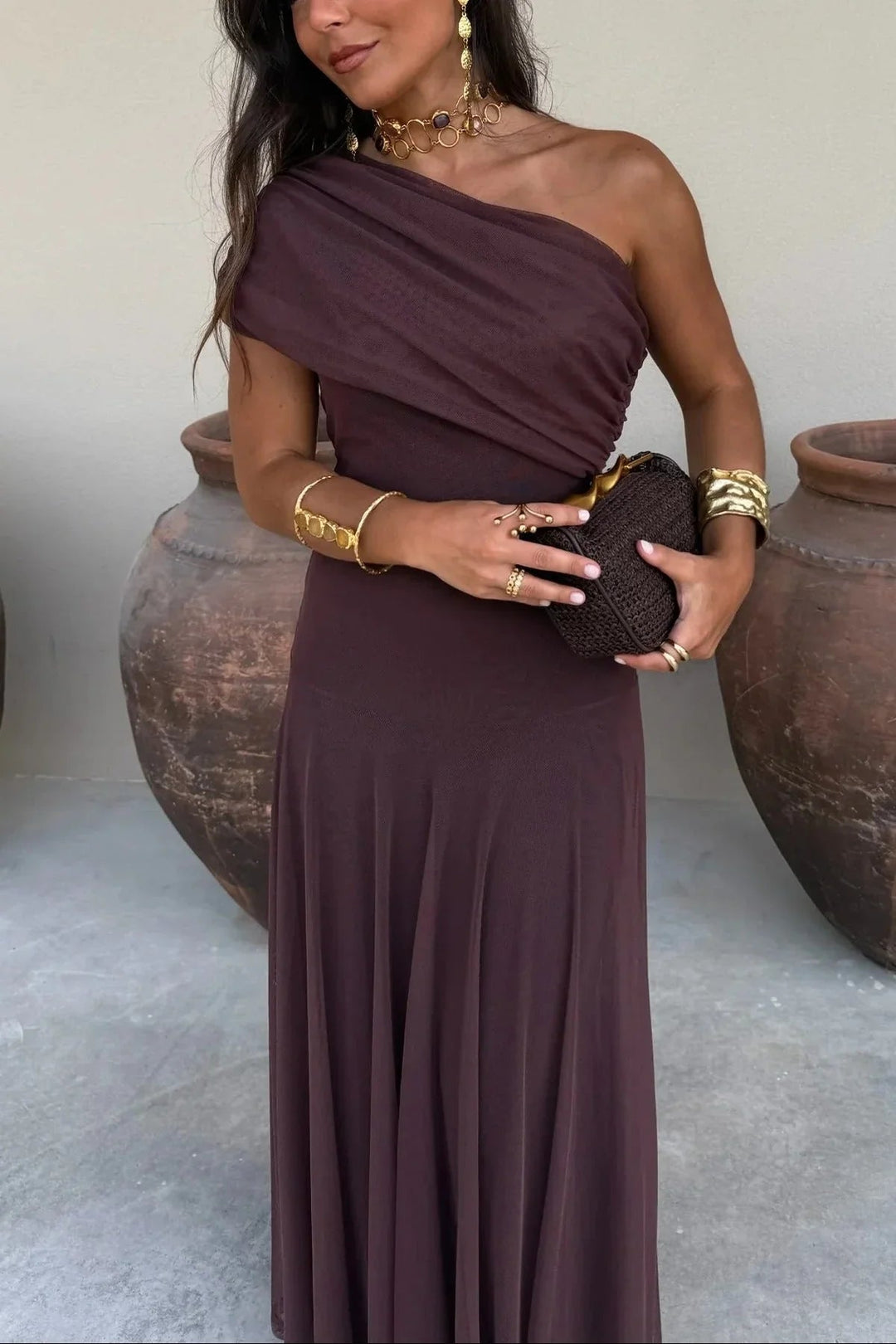 Dark brown one-shoulder maxi dress with a pleated bodice and fluid skirt, styled with gold jewelry and a woven clutch.