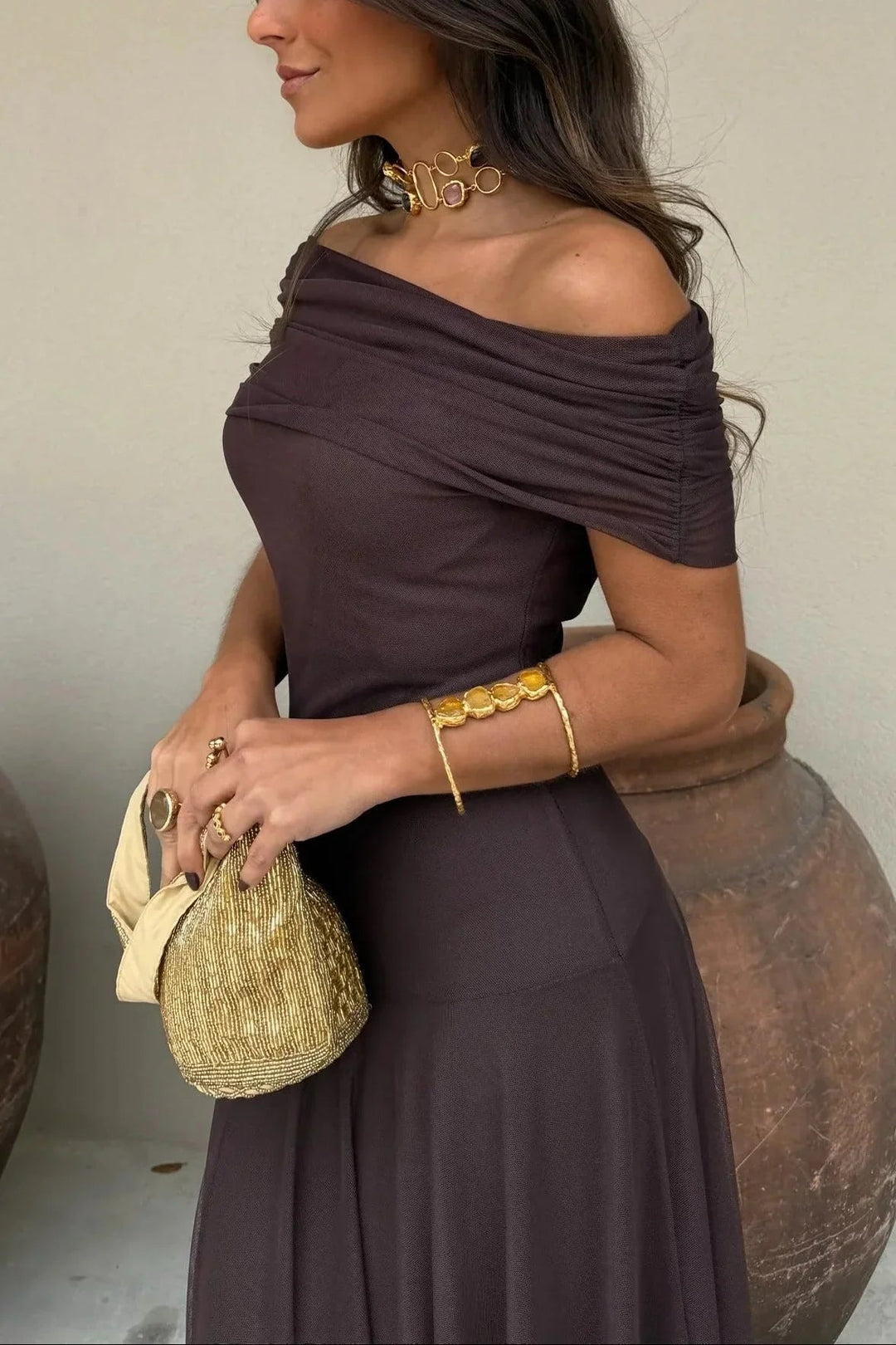 Dark brown one-shoulder maxi dress with a pleated bodice and fluid skirt, styled with gold jewelry and a beaded bag.