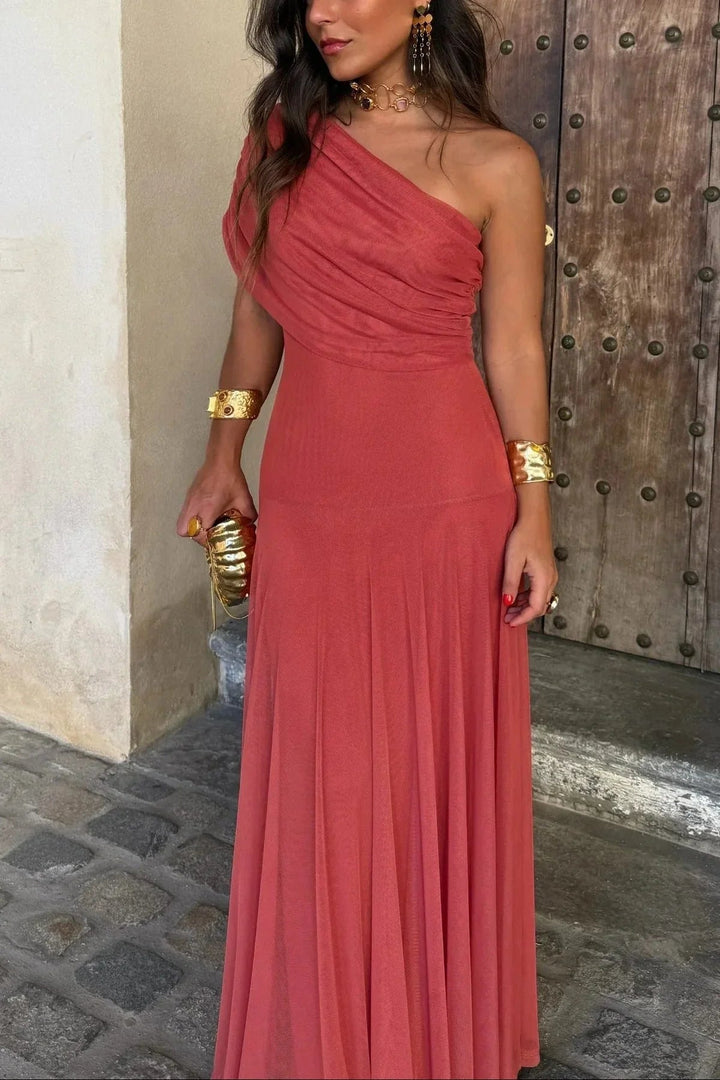 Woman wearing a terracotta one-shoulder pleated maxi dress with a fluid skirt and gold accessories.