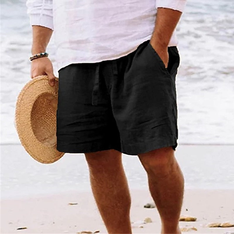 Black linen beach shorts with an elasticated drawstring waist and side pockets, worn by a man on a sandy beach.