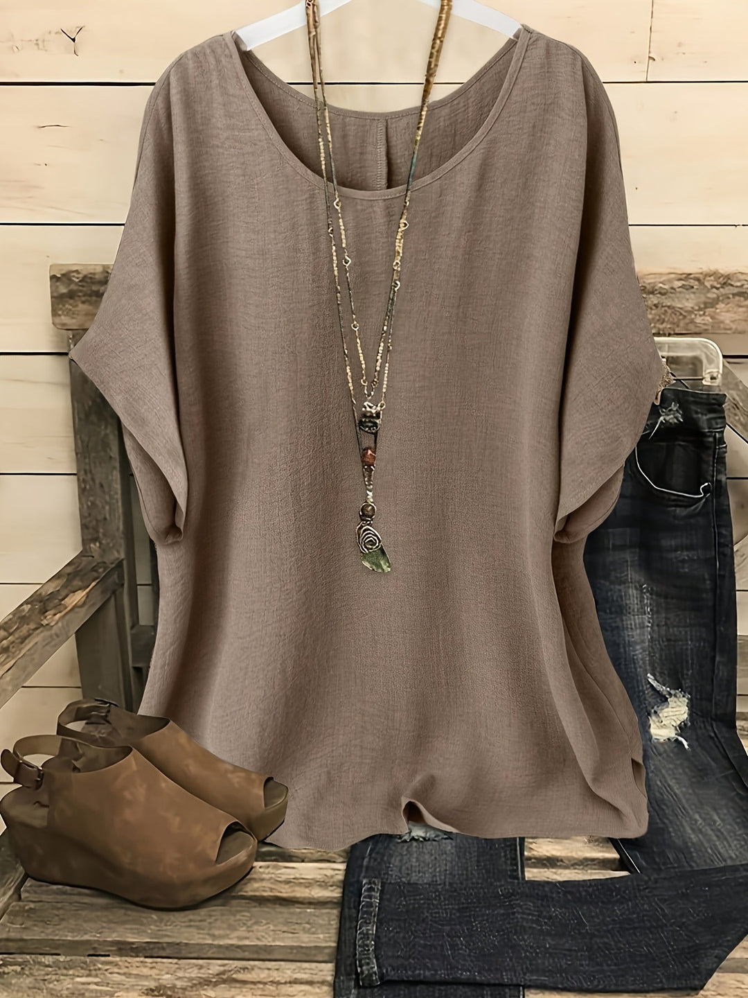 Taupe linen-look round neck top with batwing sleeves, styled with a layered necklace, distressed jeans, and wedge sandals.