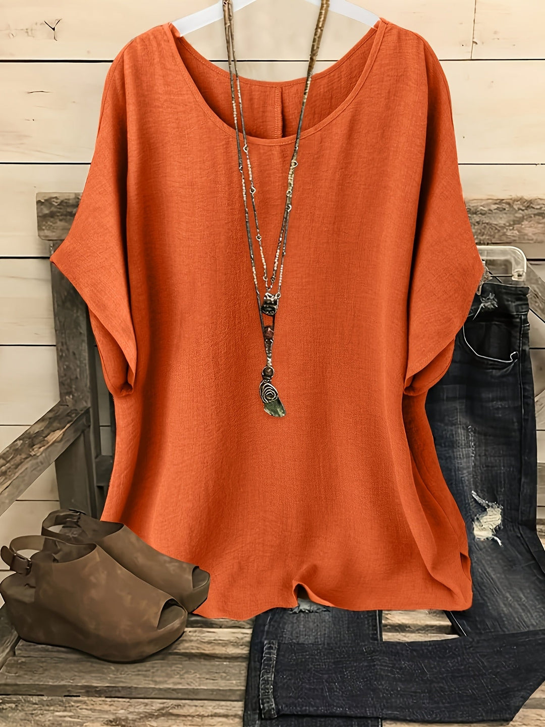 Rust orange linen-look round neck top with batwing sleeves, styled with a layered necklace, jeans, and wedge sandals.