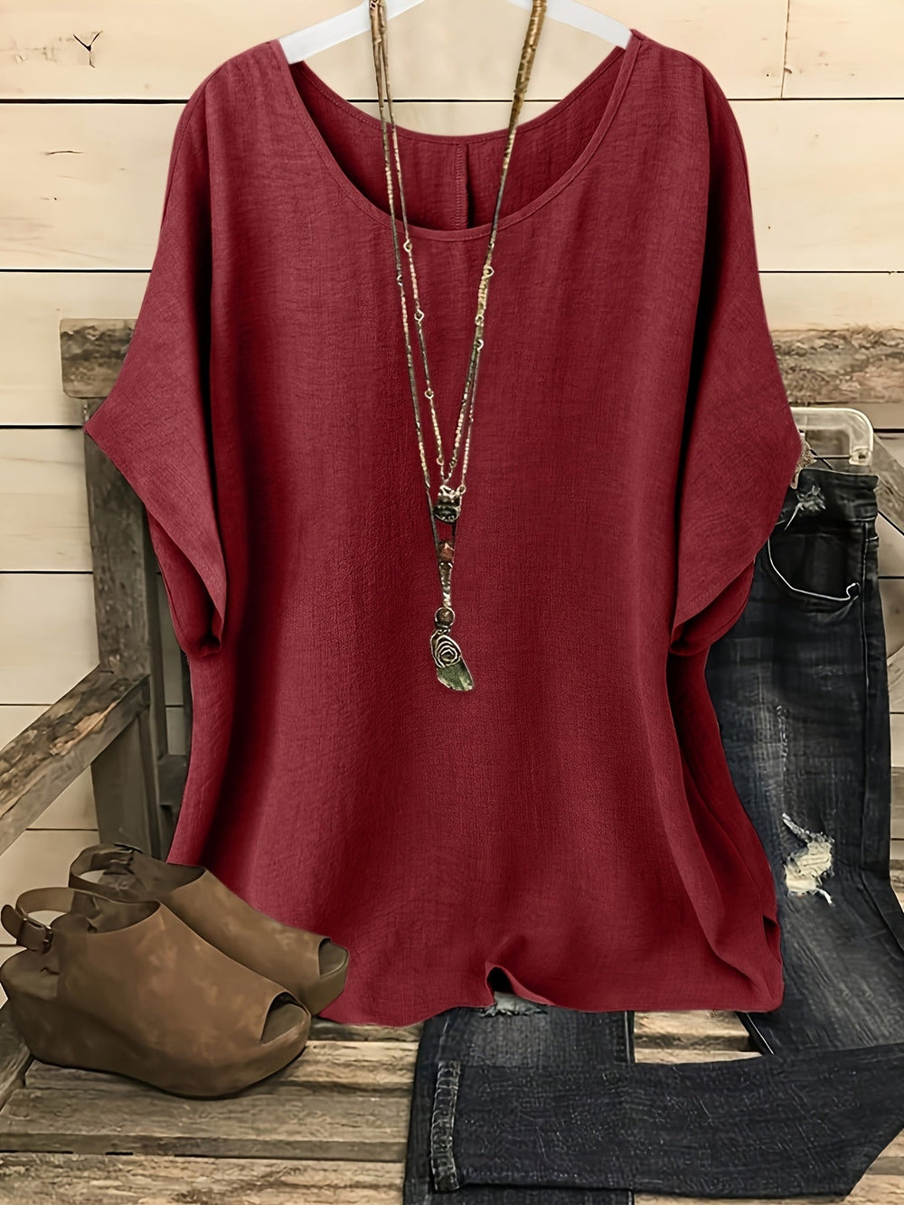 Burgundy linen-look round neck top with batwing sleeves, styled with a layered necklace, jeans, and wedge sandals.