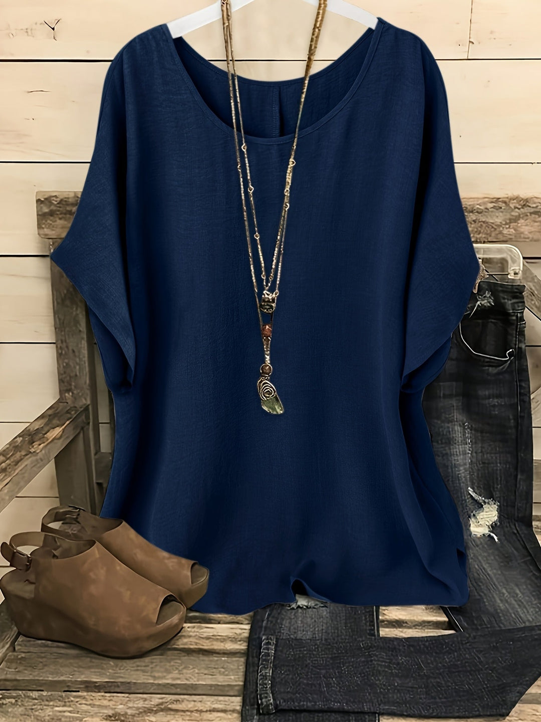 Navy blue linen-look round neck top with batwing sleeves, styled with a layered necklace, jeans, and wedge sandals.