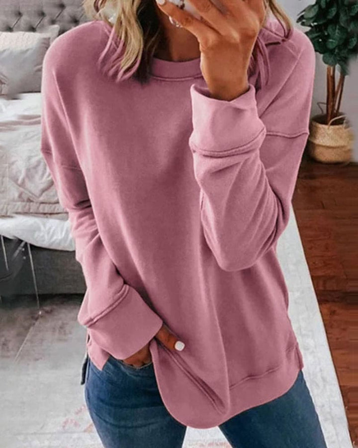 Dusty rose crew neck sweatshirt with long sleeves, ribbed cuffs, and side hem slits worn with blue jeans.