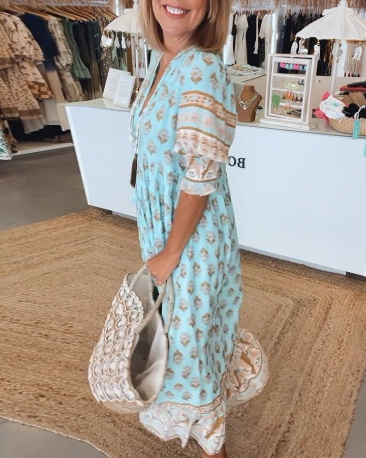 Matilda floral V-neck maxi dress in light blue with brown and white print, worn by a woman carrying a woven bag.