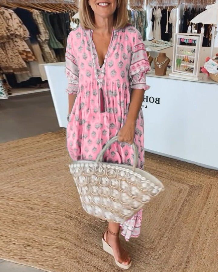 Woman in a pink floral V-neck maxi dress with a tassel tie, carrying a woven bag, in a boutique.