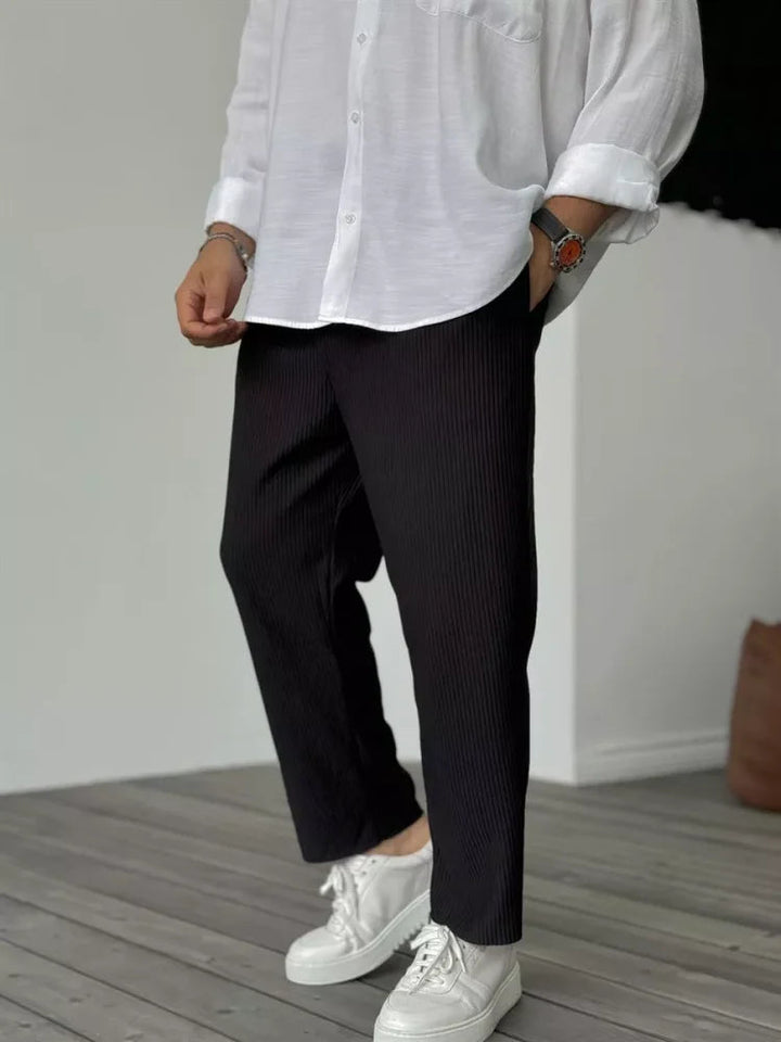 Black tapered trousers with a vertical ribbed texture, elasticated waist, and relaxed fit paired with a white shirt.