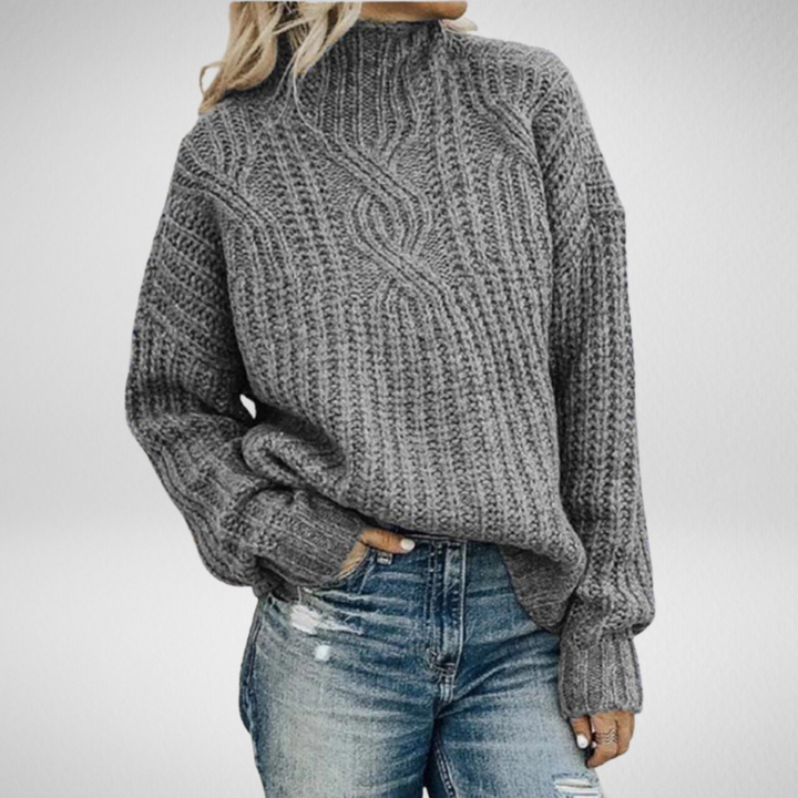 Grey chunky cable-knit jumper with a mock neck and relaxed fit, styled with distressed blue jeans.