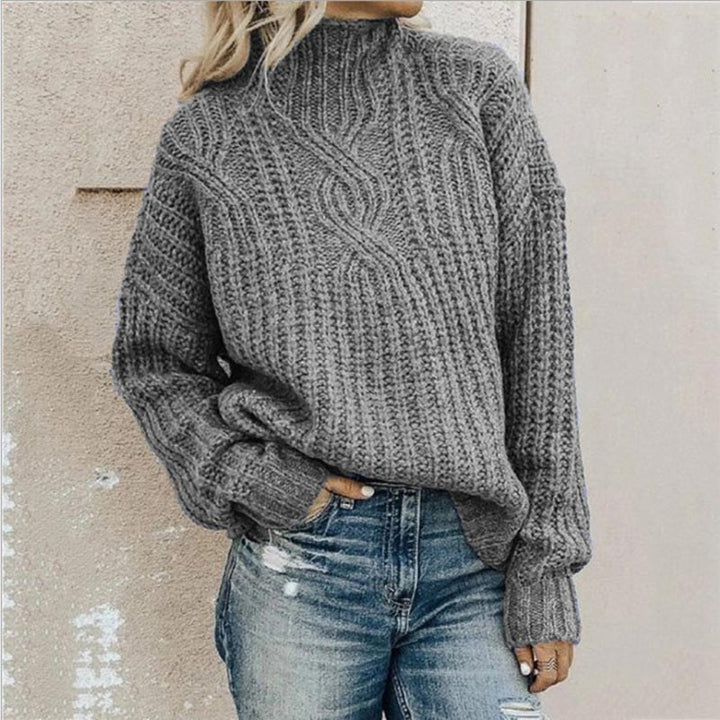 Grey cable-knit mock neck jumper with a relaxed, chunky texture and long sleeves worn with blue jeans.