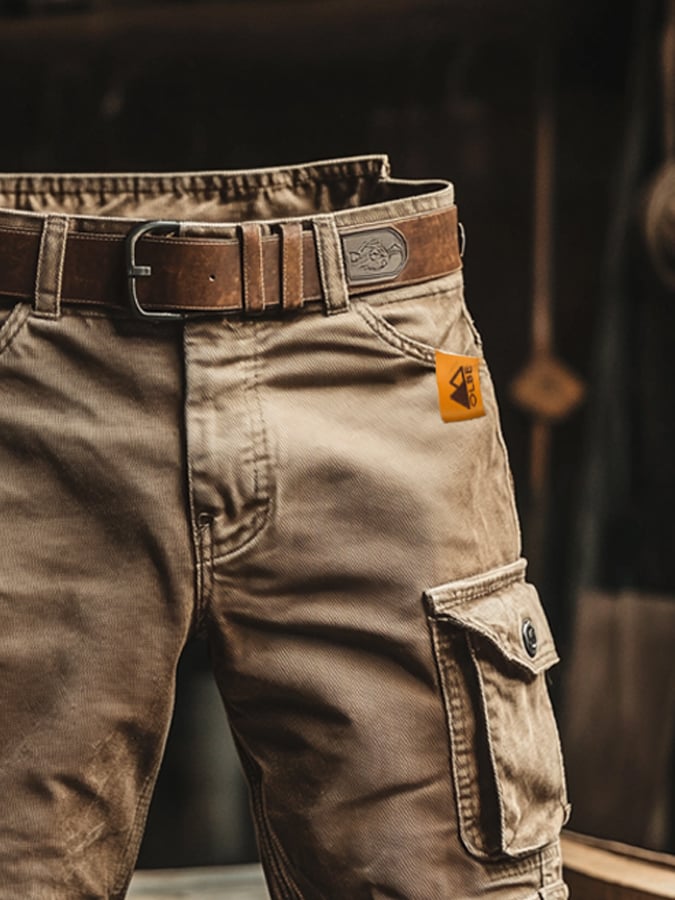 Rugged tan cargo shorts with a brown leather belt, side pockets, and an orange brand tag on a dark, rustic background.