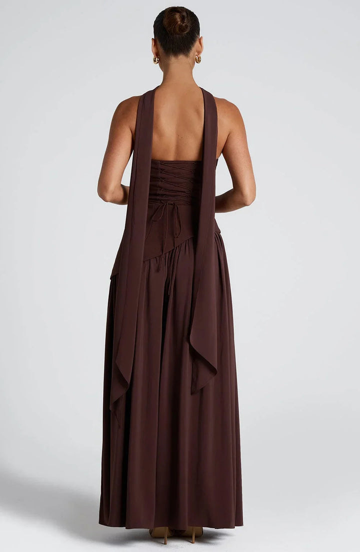Back view of a woman in a brown Phoebe corset bustier maxi dress with draped scarf detail and floor-sweeping length.
