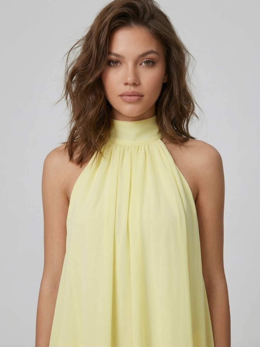 Model wearing a light yellow, sleeveless halter neck mini dress with a high neckline and airy, flowy fabric.