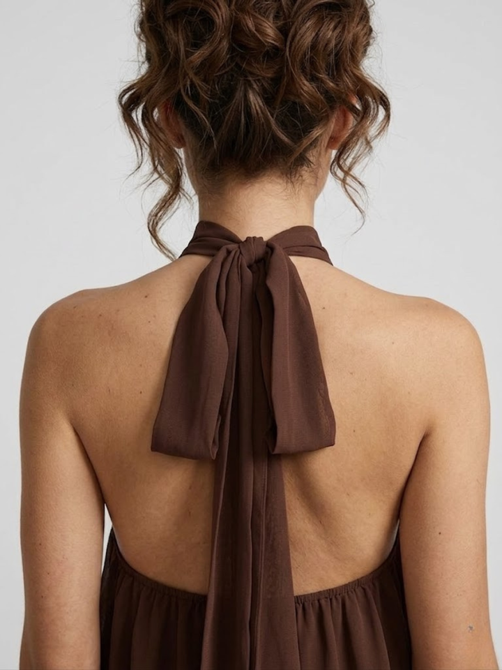 Back view of a brown halter mini dress featuring an open back and a delicate self-tie bow closure at the neck.