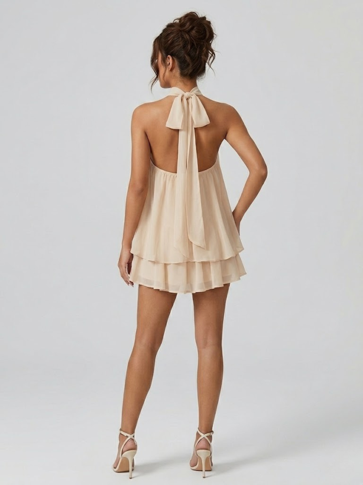 Beige backless mini dress with a halter neck, tie-back detail, and tiered ruffle hem in a lightweight, flowy fabric.