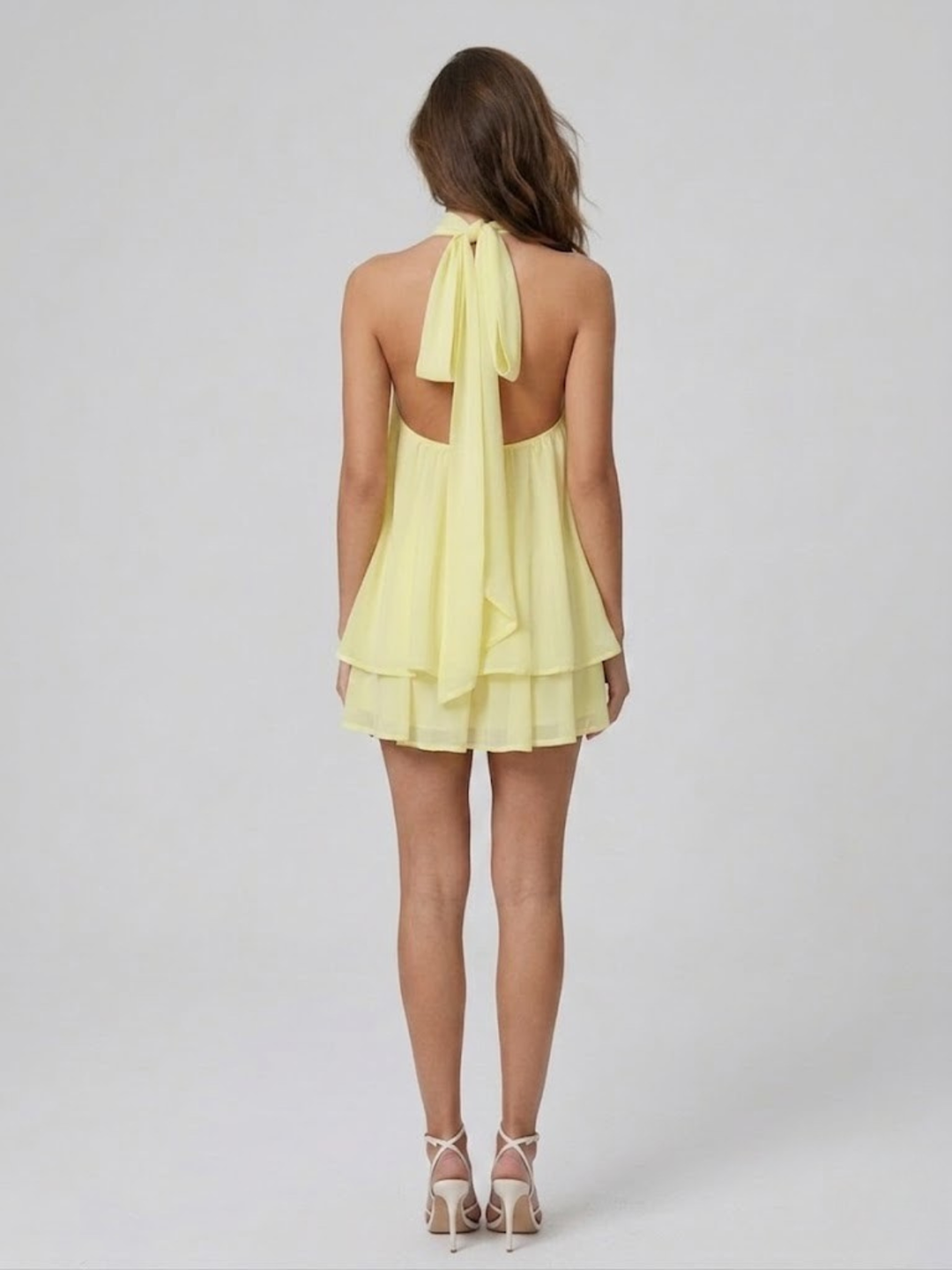 Back view of a pale yellow backless halter mini dress with a tiered ruffle hem and self-tie neck fastening.