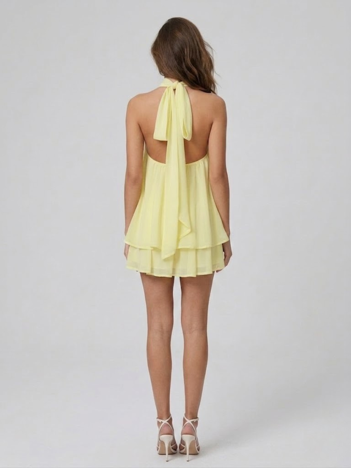 Back view of a pale yellow backless halter mini dress with a tiered ruffle hem and self-tie neck fastening.