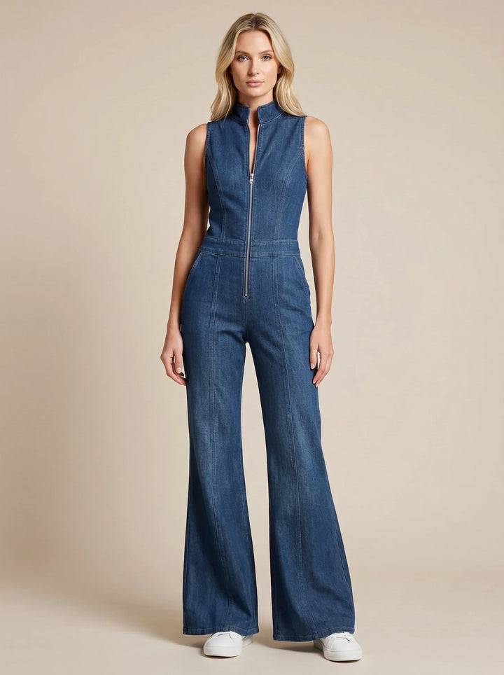 Sleeveless blue denim jumpsuit with a front zip, stand collar, fitted bodice, and flared legs worn with white trainers.