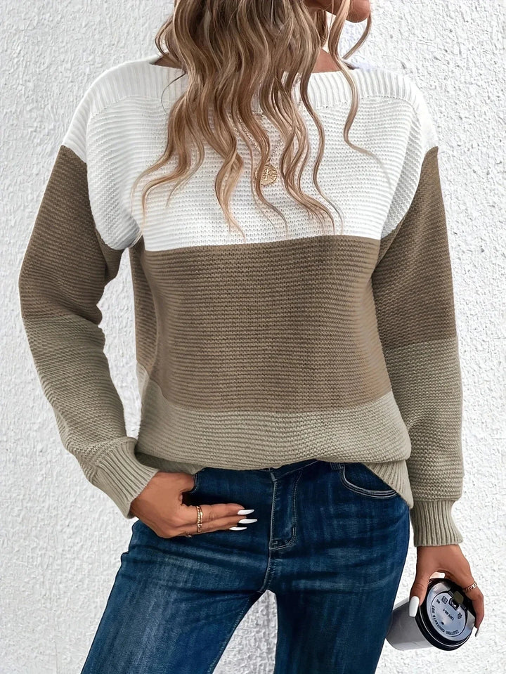 Woman wearing a chunky, ribbed colour block jumper in white, tan, and beige with a relaxed boat neckline and long sleeves.