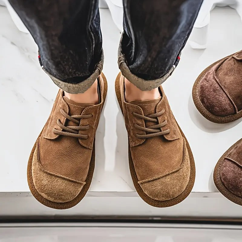 Men's brown leather lace-up walking shoes with a rounded suede toe cap and cushioned sole, worn with dark cuffed jeans.