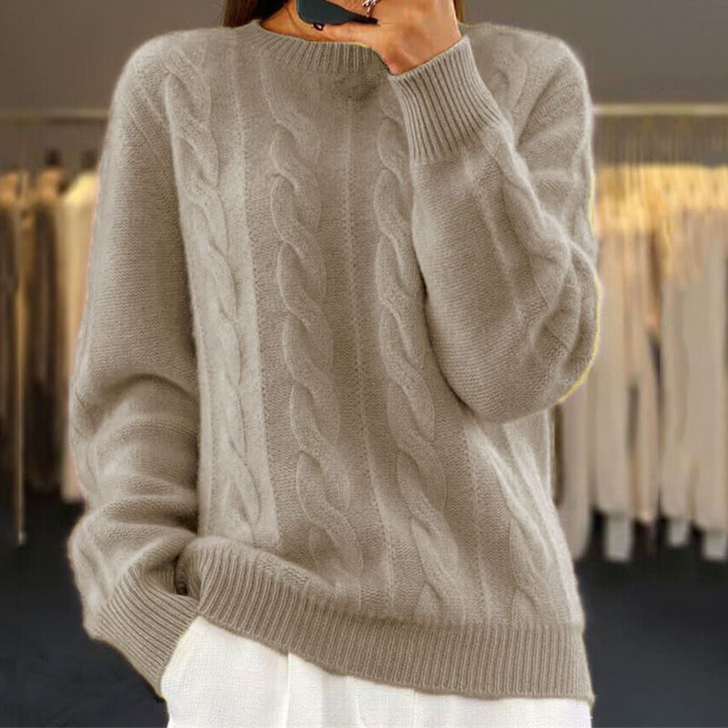 Beige cable knit crew neck jumper with a relaxed fit, ribbed cuffs, and hem, worn over white trousers.