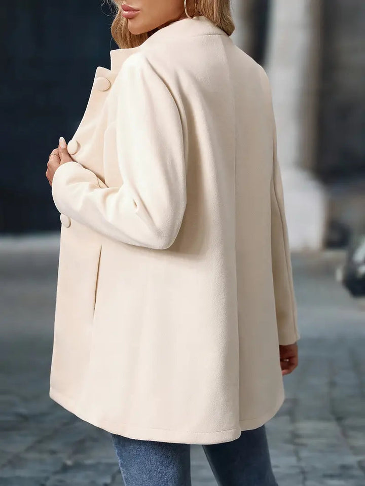 Cream-colored mid-length coat with a stand collar, button-front closure, and tailored silhouette worn over blue jeans.