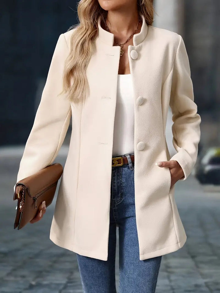 Cream mid-length coat with a stand collar, covered button front, and tailored waist, styled with jeans and a clutch.